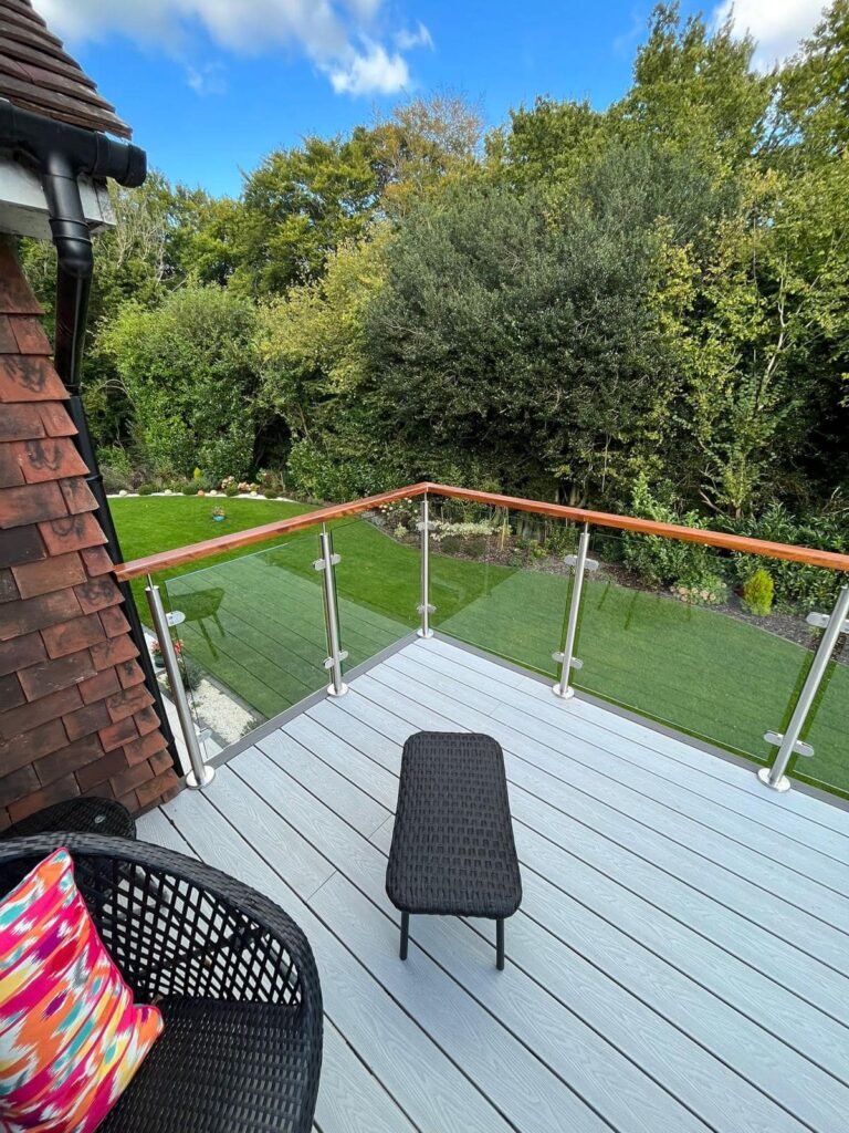 A custom-made wooden iroko balcony featuring intricate glass panel railings and an oiled natural wood finish, overlooking a scenic view. - contact us 