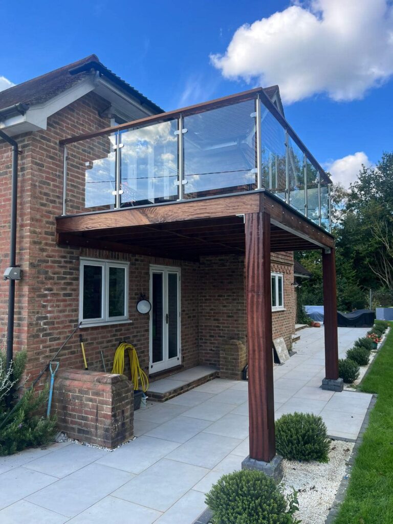 A custom-made wooden iroko balcony featuring intricate glass panel railings and an oiled natural wood finish, overlooking a scenic view. - contact us 